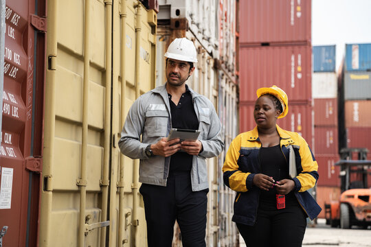 Man and woman logistics workers in high visibility safety gear discuss operations and checking container at shipping container yard	