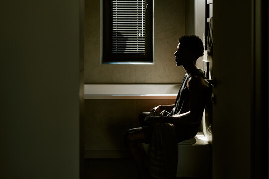 Young adult Black man sitting on bathtub edge in morning light, gazing thoughtfully toward window, short curly hair visible, relaxed posture, towel draped over lap, minimalist bathroom