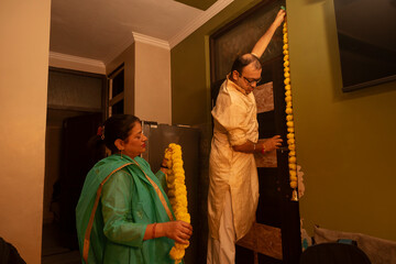 A man carefully hangs marigold garlands while his wife assists with more decorations