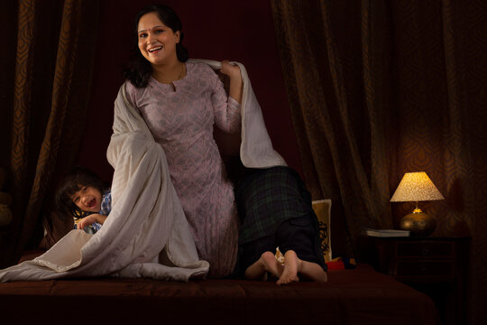Happy mother playing with her two little kids with blanket on bed