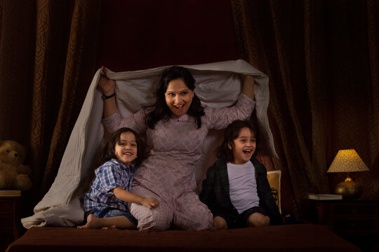 Happy mother playing with her two little kids with blanket on bed