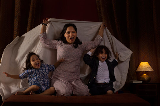 Happy mother playing with her two little kids with blanket on bed