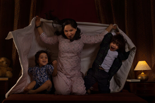 Happy mother playing with her two little kids with blanket on bed
