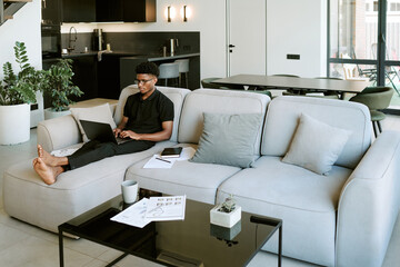 Young adult Black man sitting barefoot on sofa using laptop, working in modern living room with documents and notebook on table, morning sunlight streaming through window