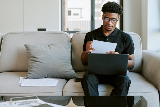 Young adult Black man sitting on sofa reviewing documents while working on laptop in modern living room, holding paper in hands, focused expression, morning light coming through window