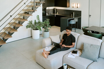 Young adult Black man sitting barefoot on sofa using laptop in modern living room, working remotely in morning, papers and notebook lying nearby, staircase and kitchen in background