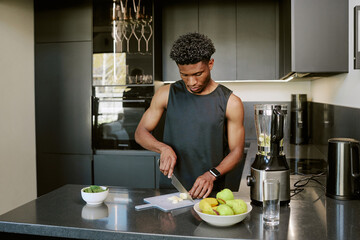 Young adult Black man preparing healthy breakfast by slicing banana on cutting board in modern...
