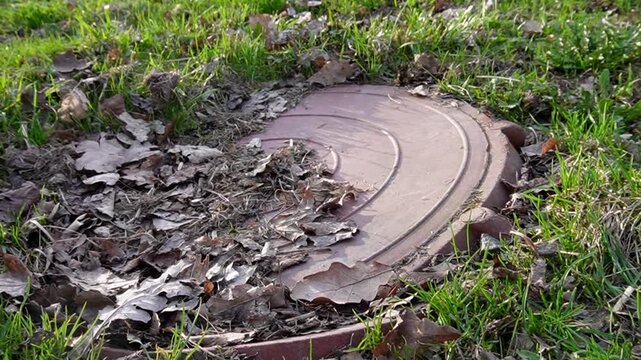 There is an old sewer manhole in the grass on the slope.In autumn,the brown manhole is at an angle, under a layer of dry leaves.The rays of setting sun fall on grass.High quality FullHD footag, video