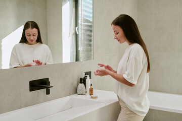 Caucasian young woman standing at bathroom sink applying skincare product to hands, looking down with relaxed expression, morning light streaming through window, mirror reflecting her