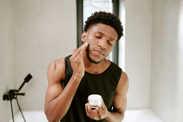 Young adult Black man standing in bathroom applying facial cream to cheek with eyes closed, holding skincare jar in hand, morning beauty routine emphasizing self care and grooming