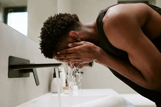 Young adult Black man washing face at bathroom sink in morning, leaning forward with hands cupping water, short curly hair visible, starting daily skincare routine