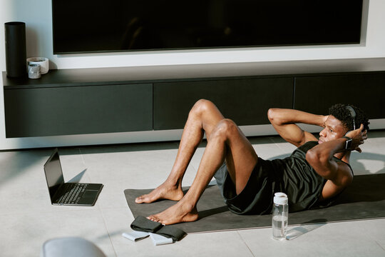 Young adult Black man exercising on yoga mat performing crunches while watching workout video on laptop in modern living room, headphones on, water bottle and towel nearby