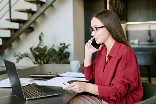 Caucasian young adult woman sitting at desk talking on smartphone while working on laptop, holding documents with charts, wearing eyeglasses, focused on multitasking in modern home office