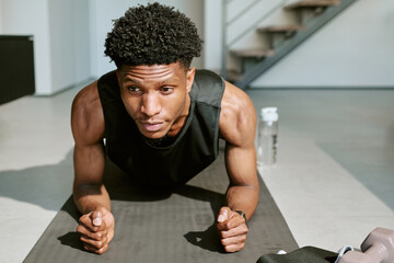 Portrait of young adult Black man exercising on yoga mat, holding plank position indoors during morning, focused expression, fitness equipment and water bottle visible nearby