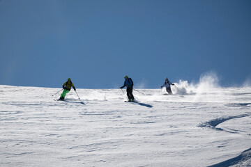Three skiers carve down a sunny, powder-covered mountain slope, kicking up fresh snow under a clear...