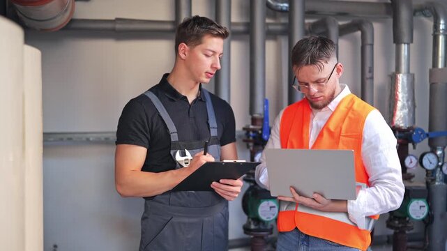 Professional engineers diligently review industrial system data, one recording notes on clipboard, other analyzing information on laptop. Focused, collaborative work, teamwork and technical expertise.