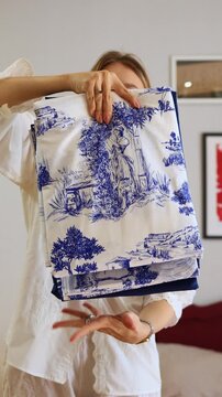 A young woman holding up and presenting newly unpacked dark blue and white Toile de Jouy patterned bed linen, focusing on the beautiful textile design and the quality of the new home goods
