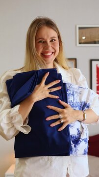 A cheerful young woman in a white blouse happily holding folded, new bed linen &ndash; solid dark blue and white Toile de Jouy pattern &ndash; promoting domestic tasks, fresh bedding, and home care.