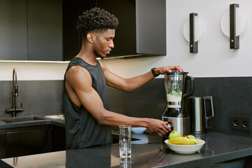 Young adult Black man preparing smoothie using blender in modern kitchen, standing at counter with fresh fruit nearby, focusing on blending ingredients for healthy morning routine