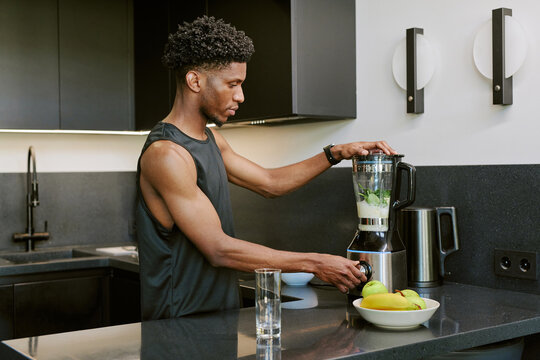 Young adult Black man preparing smoothie using blender in modern kitchen, standing at counter with fresh fruit nearby, focusing on blending ingredients for healthy morning routine