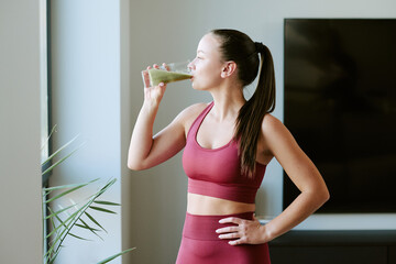 Young adult Caucasian woman standing near window drinking green smoothie with eyes closed, wearing sportswear, hand on hip, enjoying healthy morning routine after workout