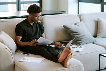 Young adult Black man sitting on sofa working on laptop and analyzing documents in bright living...