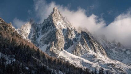 Snowy jagged mountain peaks rise above a forested slope, with clouds drifting around the snow-covered summit. Concept Snowy jagged mountain peaks, Forested slope below