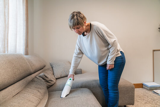 Woman cleaning sofa using a cordless handheld vacuum cleaner in a modern living room. Keeping the home tidy and dust free