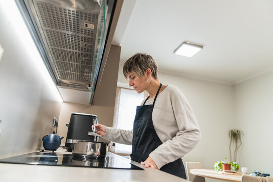Woman in apron cooking on an induction hob in a bright modern kitchen, stirring a saucepan domestic meal prep and focused home life
