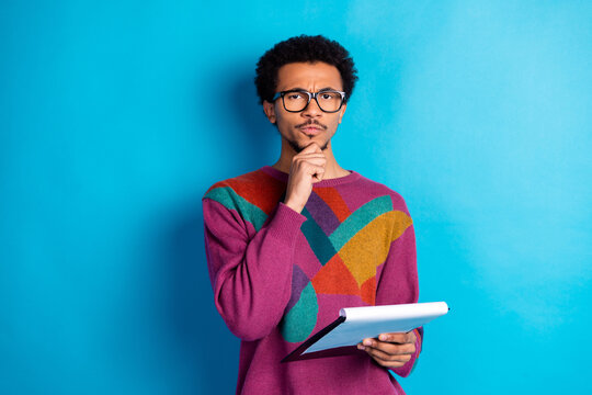 Young man in colorful sweater holding a notepad and thinking against a blue background - Powered by Adobe