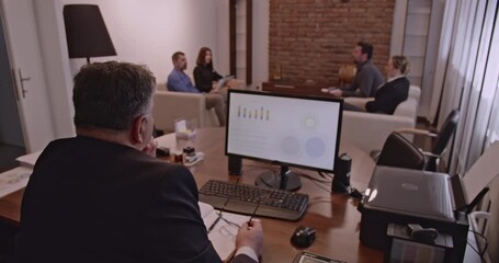 Business people in a meeting discussing data and analytics. A businessman sits at his desk while a group of colleagues meet on a sofa. Business meeting in modern office. - Powered by Adobe