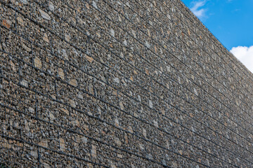 Large gabion retaining wall filled with stones, built for slope protection and erosion control, photographed against a blue sky.