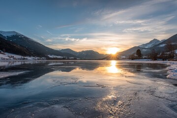Frozen lake reflecting golden sunset sunbeams on snow covered mountains and blue sky winter
