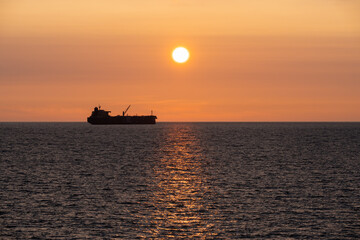 Fototapeta premium Silhouette of a large industrial tanker ship on the Baltic Sea horizon against a golden sunset sky and reflecting water.