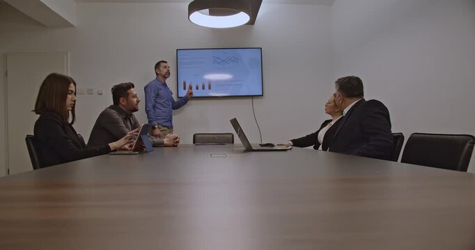 Businesspeople in a meeting discuss charts and data displayed on a monitor. A diverse team collaborates on a project in a modern office. - Powered by Adobe
