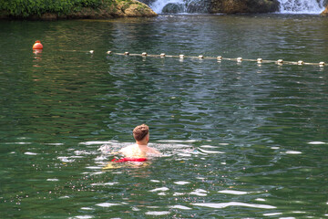 A man swimming in a clear green river near a small waterfall on a sunny day, surrounded by calm water and nature.