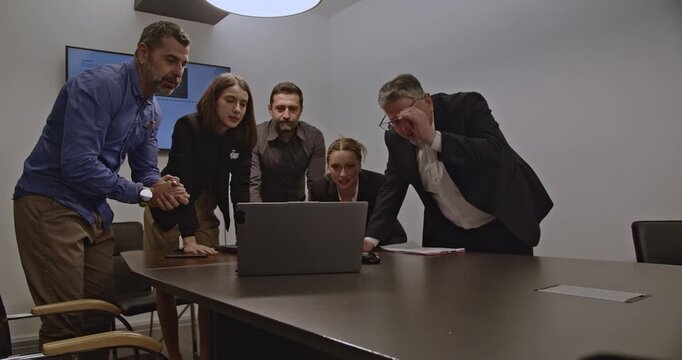 Businesspeople gather around a laptop, discussing, pointing, and expressing concern and frustration in a meeting. The atmosphere is serious, suggesting a challenging situation.