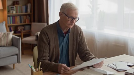 Medium shot of elderly Caucasian man in cozy room sorting through envelopes and detailed statements while organizing household expenses