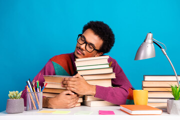 Funny Young man with trendy sweater holding stack of books smiling at desk with blue background