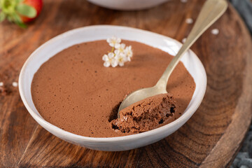 Smooth chocolate mousse in a white bowl, a golden spoon lifting a bite, garnished with tiny white flowers, presented on a textured wooden board