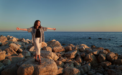 A girl poses and relaxes by the sea, standing on rocks near the slope of a high cliff, panoramic view of the seashore and mountains at sunset