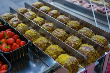 Rows of chocolate-coated dessert balls garnished with pistachio and phyllo pastry, displayed in a metal case at a street food market, fresh strawberries on the side, concept of sweet treat, street