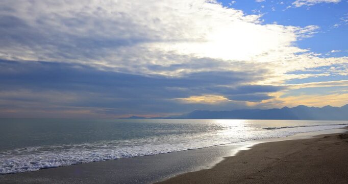 Gentle waves washing sandy shore with sunbeam breakthrough across wide horizon and distant mountain silhouettes at Sunrise Beach near Patara in Antalya Province during calm light