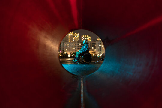 Pondering disabled young man in wheelchair sitting alone outdoors in neon rgb light, enjoying fresh air, engaged in deep thinking, reflecting on thoughts and feelings, relieving daily stress outside.