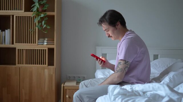 Sleepy morning of groggy man sits on bed, looks at smartphone slowly wakes up while scrolling through notifications, early fatigue. Morning procrastination of male daily routine of digital dependence.