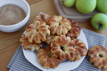Homemade puff pastry apple rings with cinnamon and cane sugar on white ceramic plate on wooden table with fresh apples