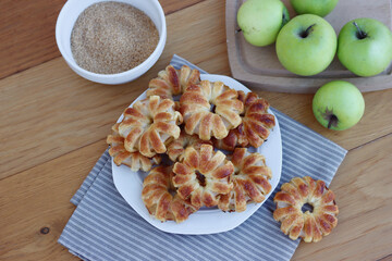 Homemade puff pastry apple rings with cinnamon and cane sugar on white ceramic plate on wooden table with fresh apples