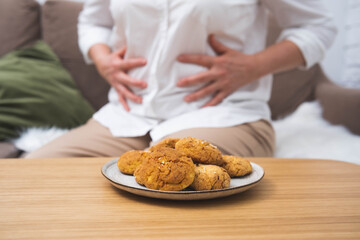 A plate of cookies in the foreground with a woman holding her stomach in discomfort in the background. Concept of overeating, food intolerance, stomach pain, or digestive issues.