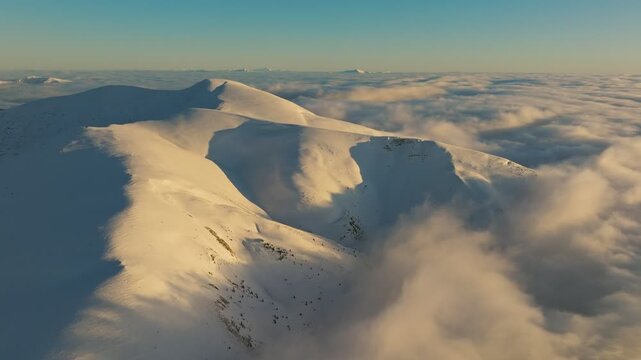 Sunlit mountain crest, Warm glow on snowy slopes, Soft clouds beneath illuminated mountain ridge, Golden evening light highlights snow contours on gentle slopes