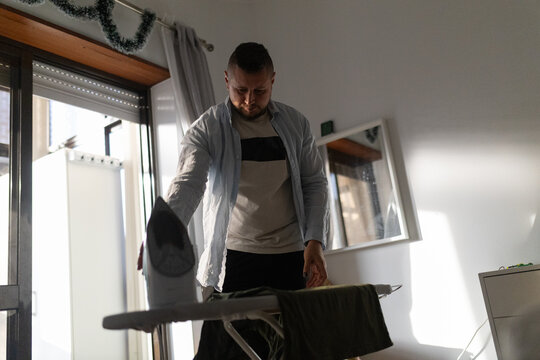 Young man ironing clothes at home in sunlight